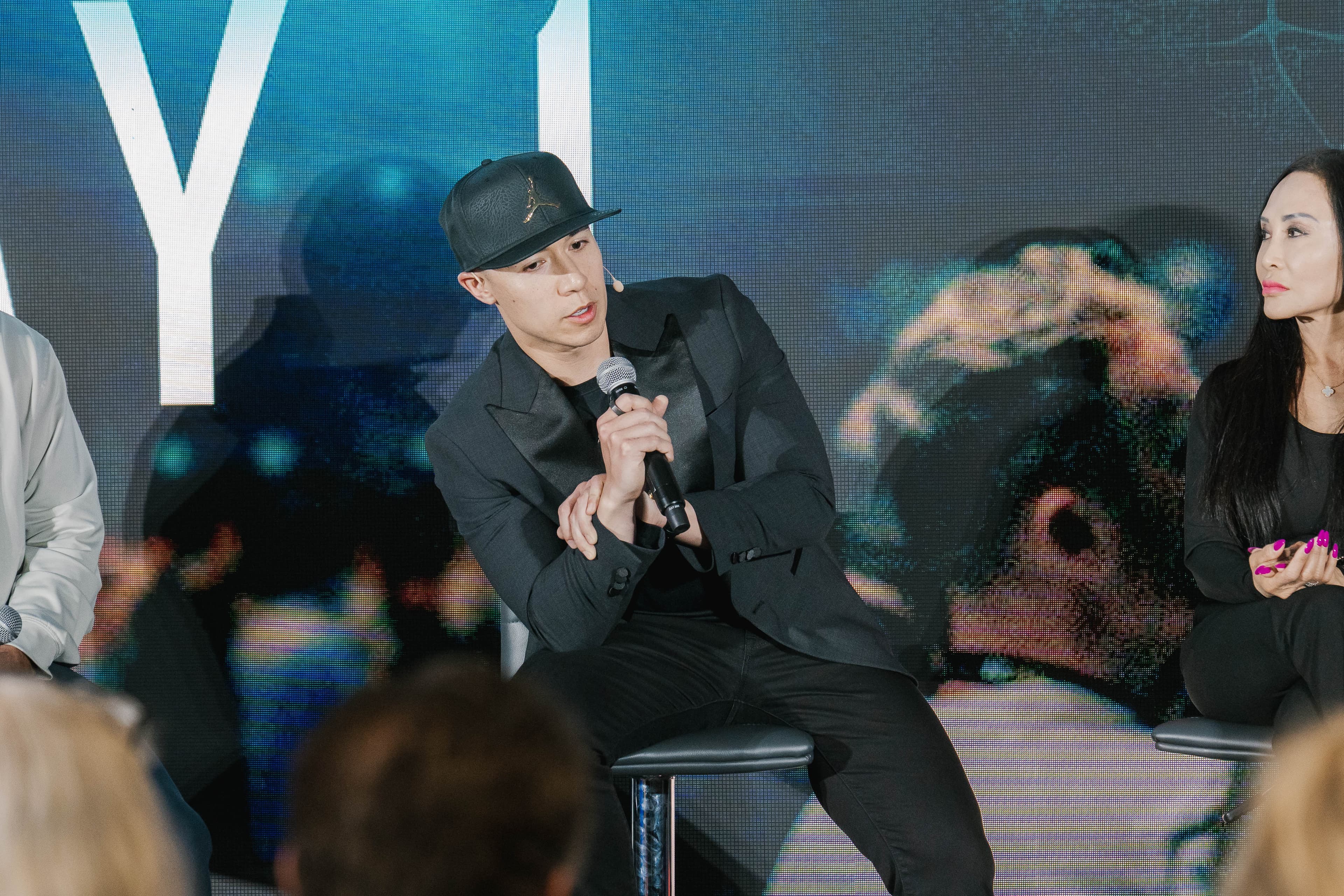 Joe Wanner on stage in a black jacket and cap, leaning forward on a stool with a microphone in hand.